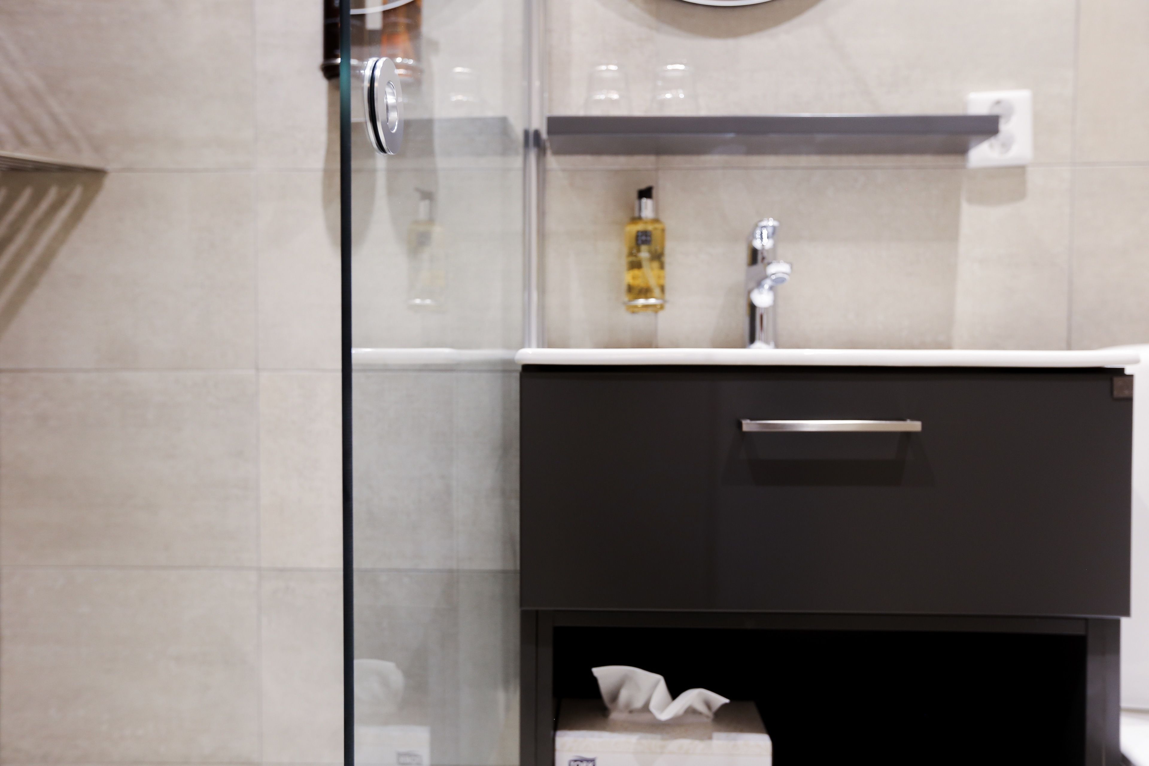 Un baño moderno con cabina de ducha de vidrio, tocador negro con grifo plateado y una pequeña estantería con artículos de aseo.