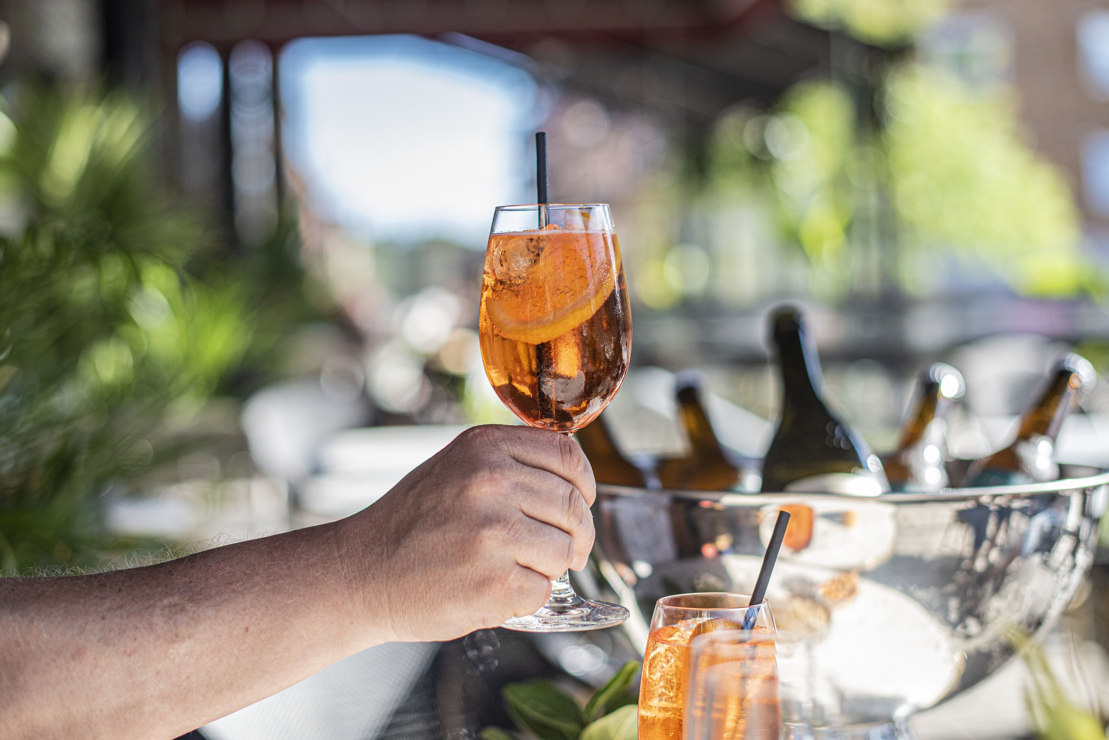 Eine Hand hält ein Glas mit einem Getränk, Eiswürfeln und einer Zitronenscheibe. Im Hintergrund steht eine Champagnerflasche.