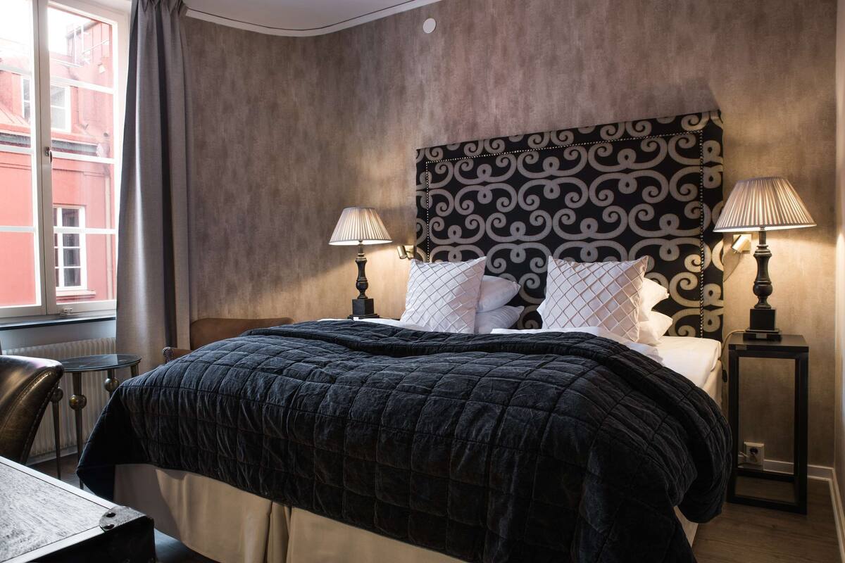 Une chambre d’hôtel avec un grand lit, deux lampes de chevet, une tête de lit ornée d’un motif décoratif et une vue sur un bâtiment à l’extérieur.