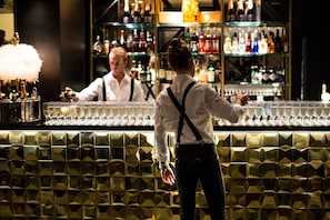Una zona de bar con un cantinero y un cliente. La barra tiene un revestimiento con un diseño y estantes llenos de botellas.