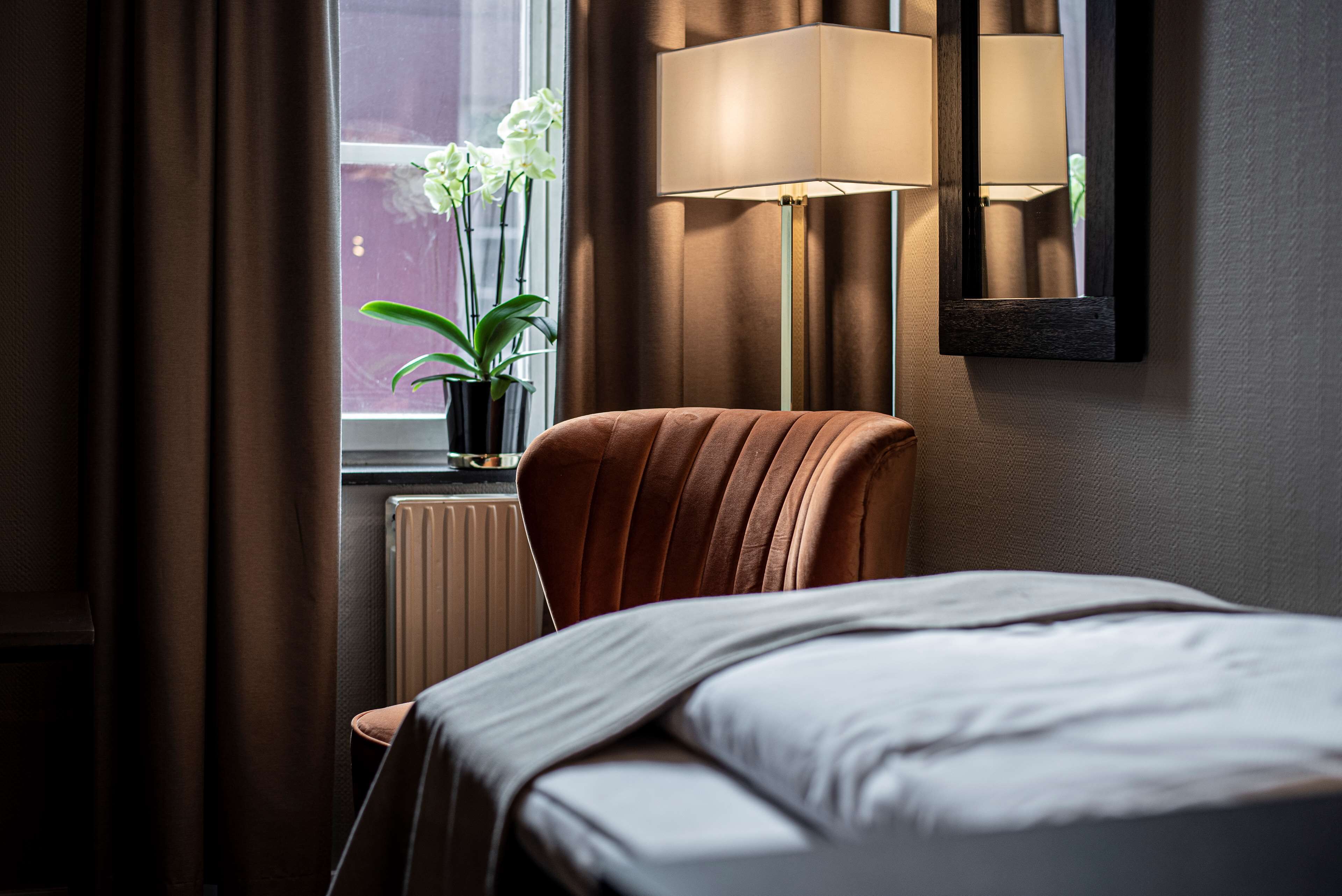 Habitación de hotel con cama, una silla, lámpara y ventana con vistas a un edificio y una planta.