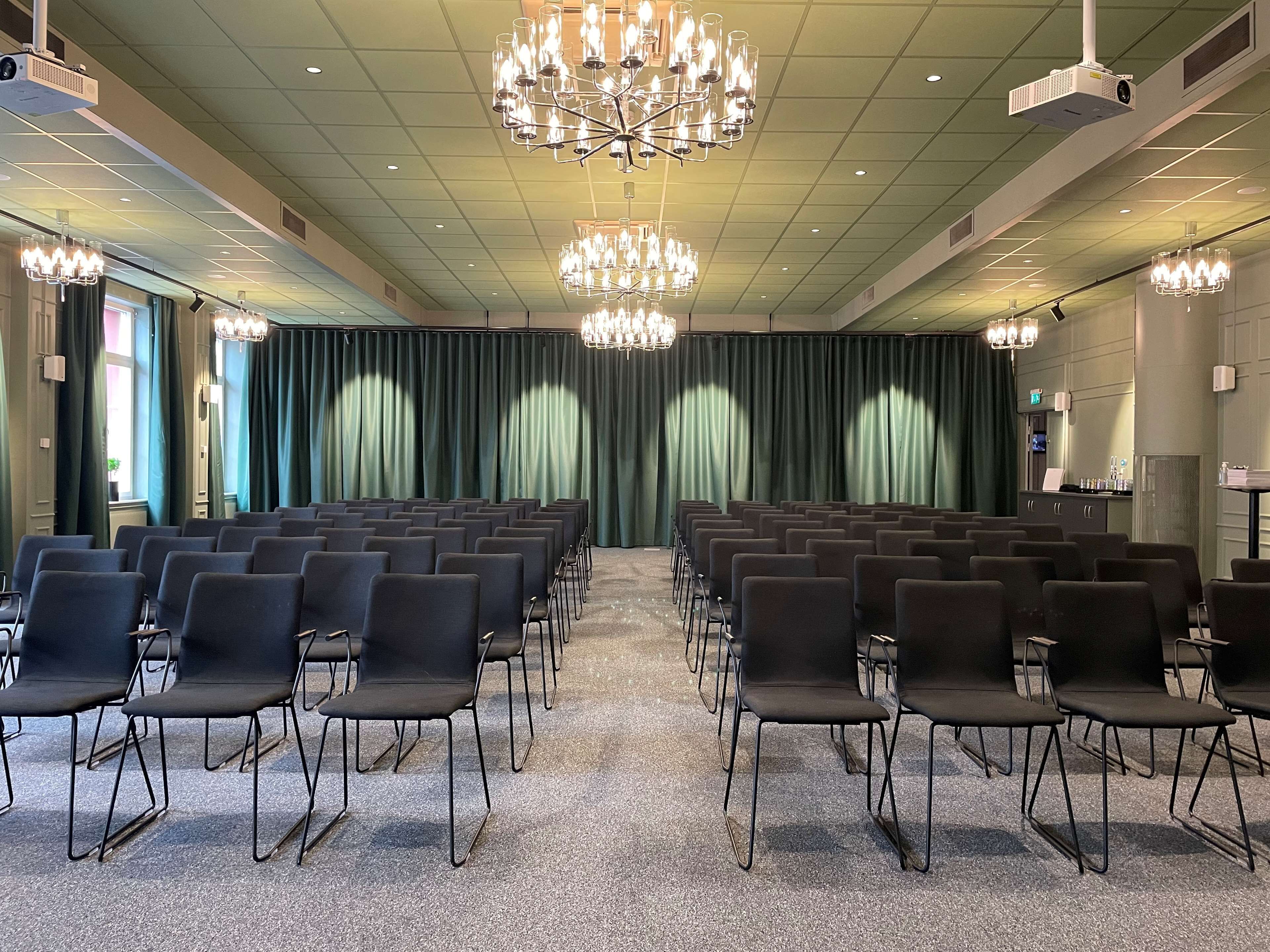 A conference room with rows of chairs, a large chandelier, and a projector on the ceiling.