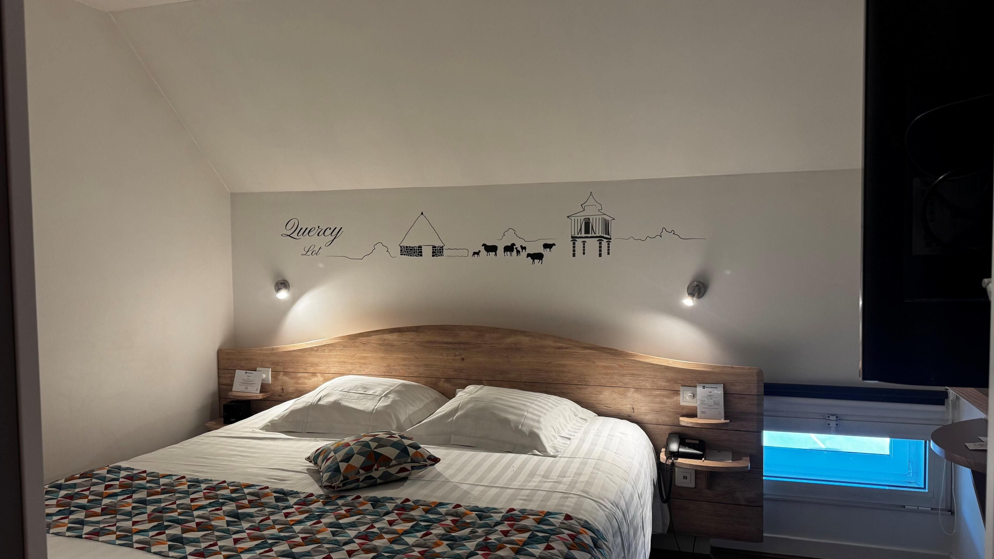 A bedroom with a wooden headboard, a bed with white linens, a colorful quilt, a wall decal with farm animals, and a skylight.