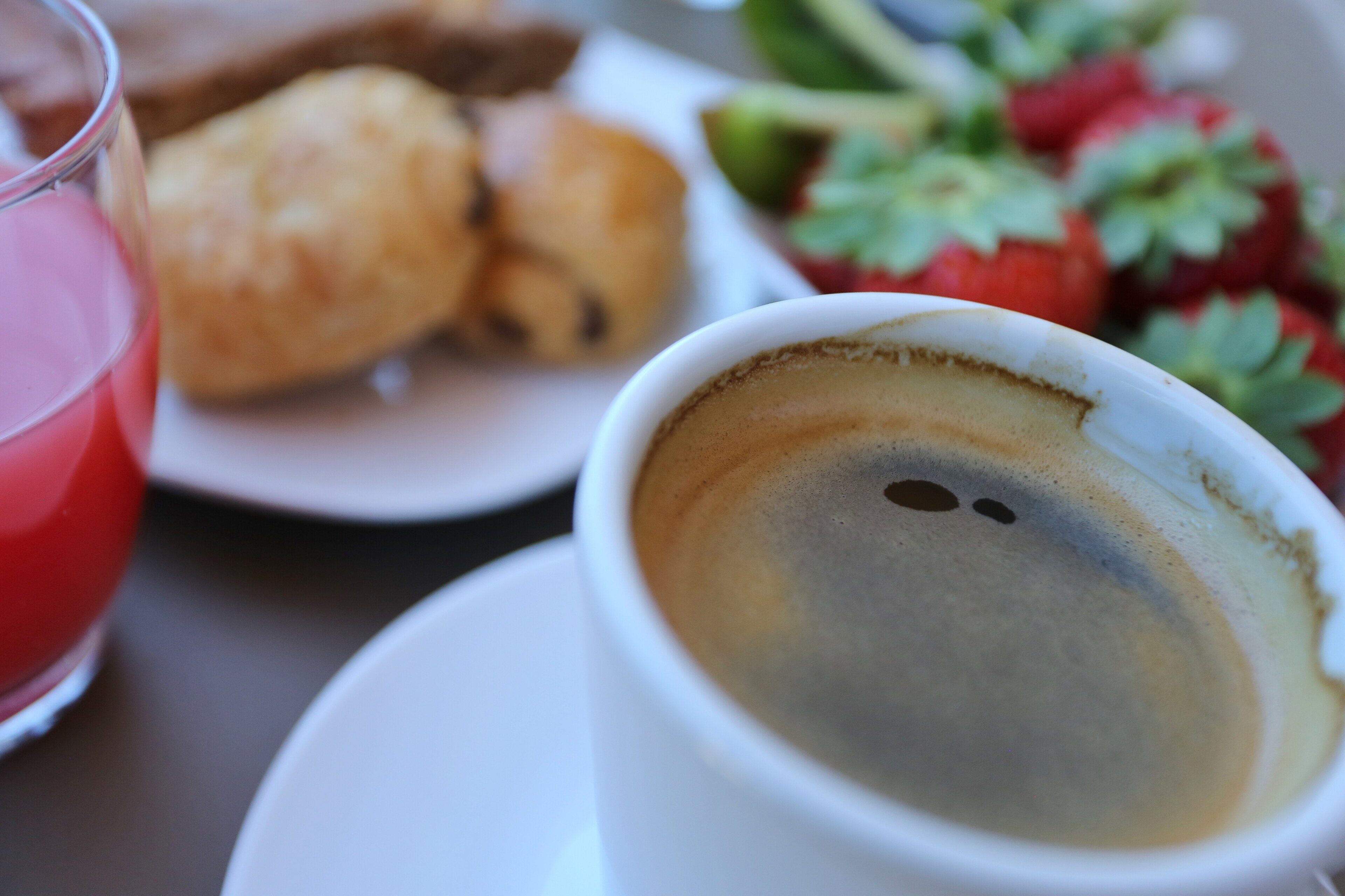 Una tassa de cafè sobre un calze, amb un got de beguda vermella a prop, i un plat amb pastissos i maduixes al fons.