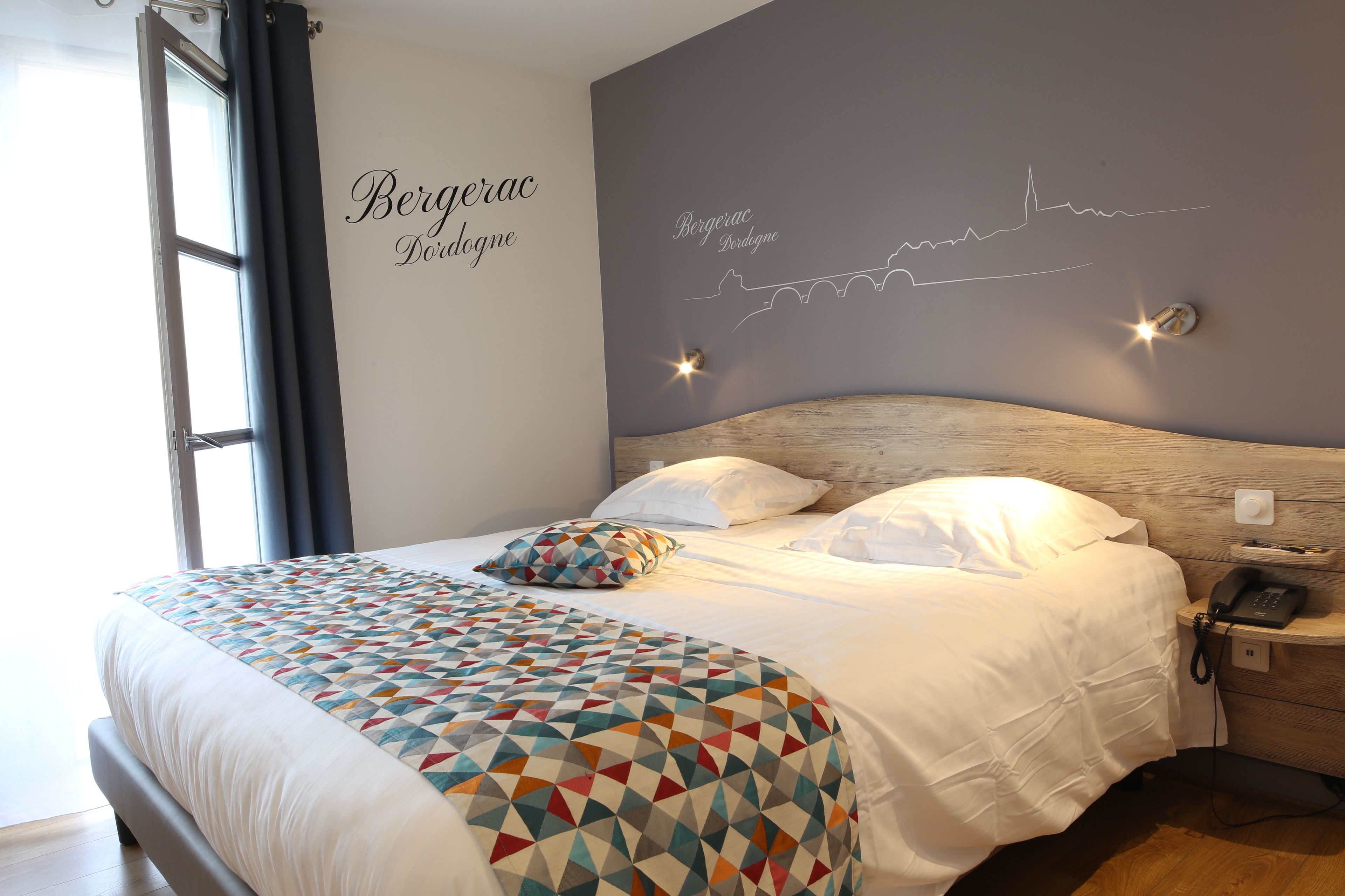 Una cama bien tendida con un edredón estampado, un teléfono en la mesita de noche y una pared con el silueta del horizonte de una ciudad.