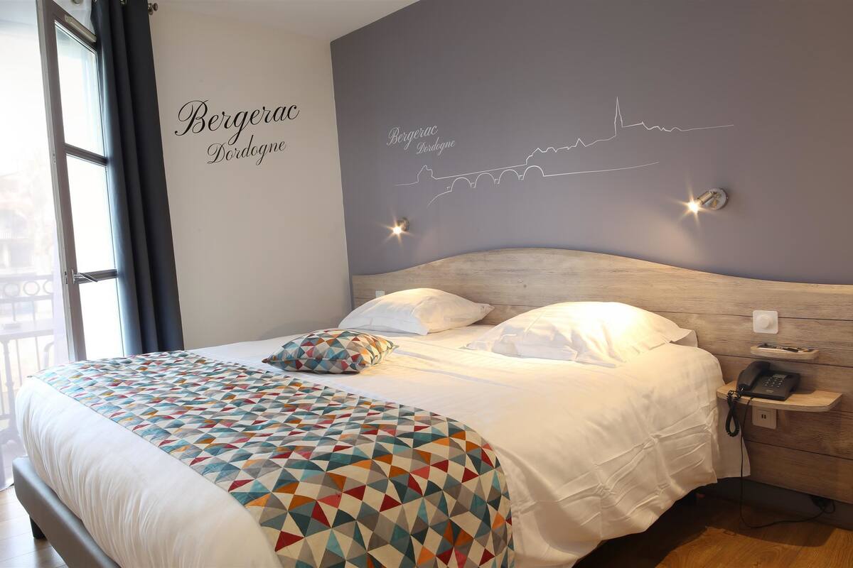 Een opgemaakt bed met een geborduurd plaid, een telefoon op het nachtkastje, en een muur met een silhouet van een stadsgezicht.