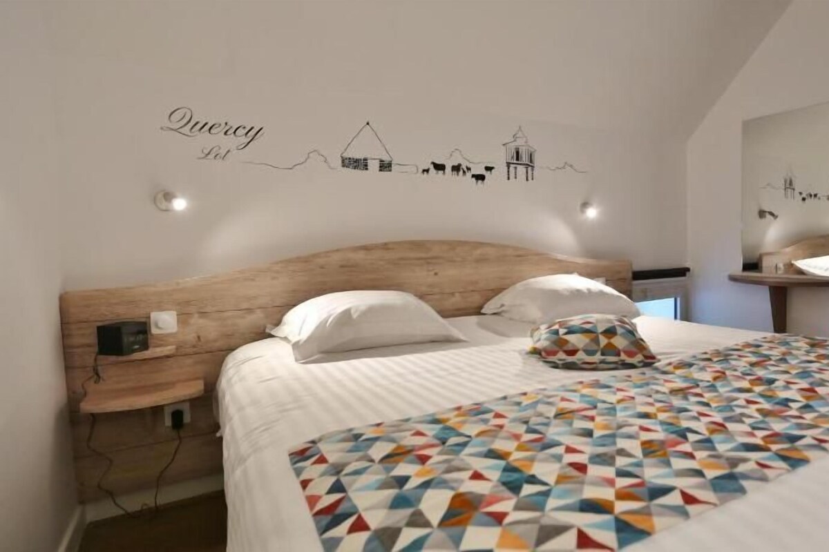 Un dormitorio con cabecera de madera, colcha estampada y un adhesivo de pared con un paisaje rural y la palabra 'Quercy'.