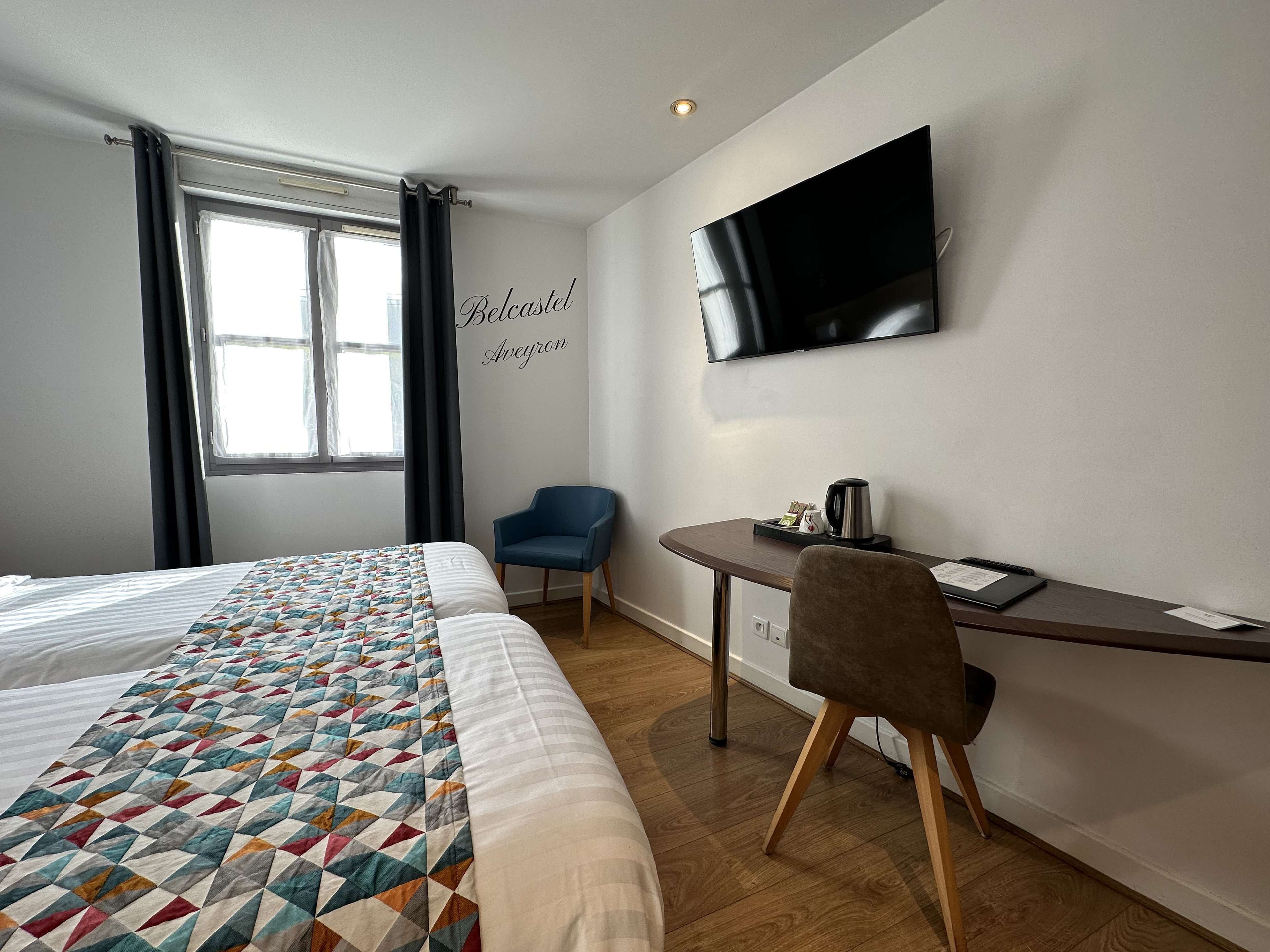 A hotel room with two beds, a desk with a chair, a flat-screen TV, and a window with curtains.