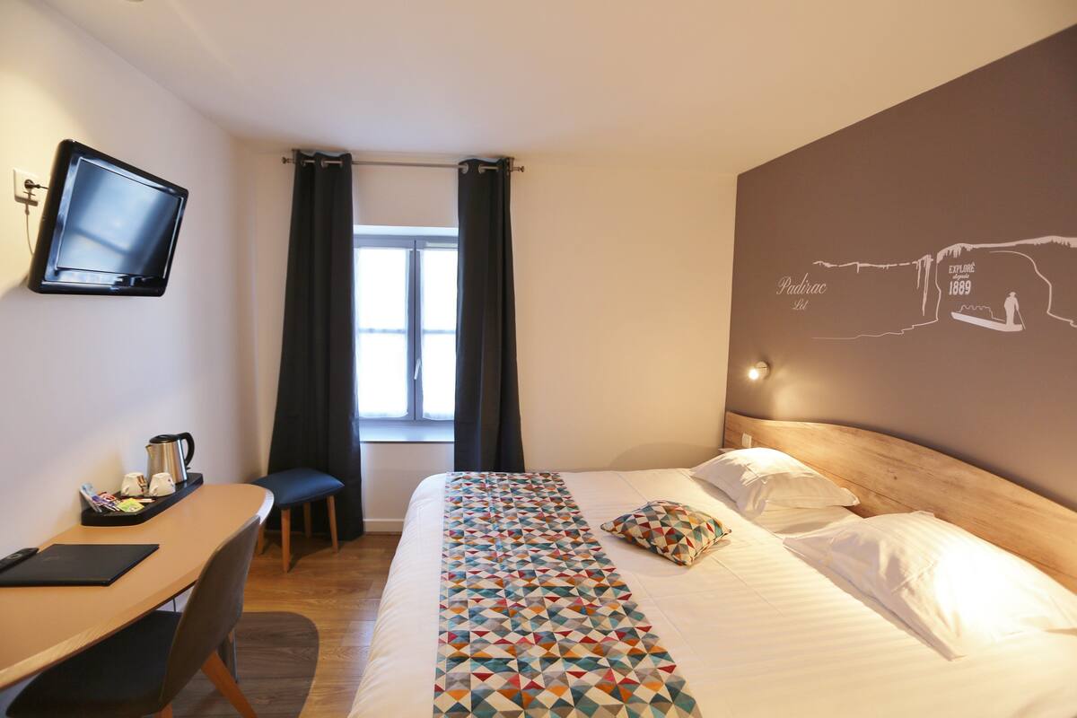 Een hotelkamer met een bed, een bureau met stoel, een televisie en een raam met gordijnen.