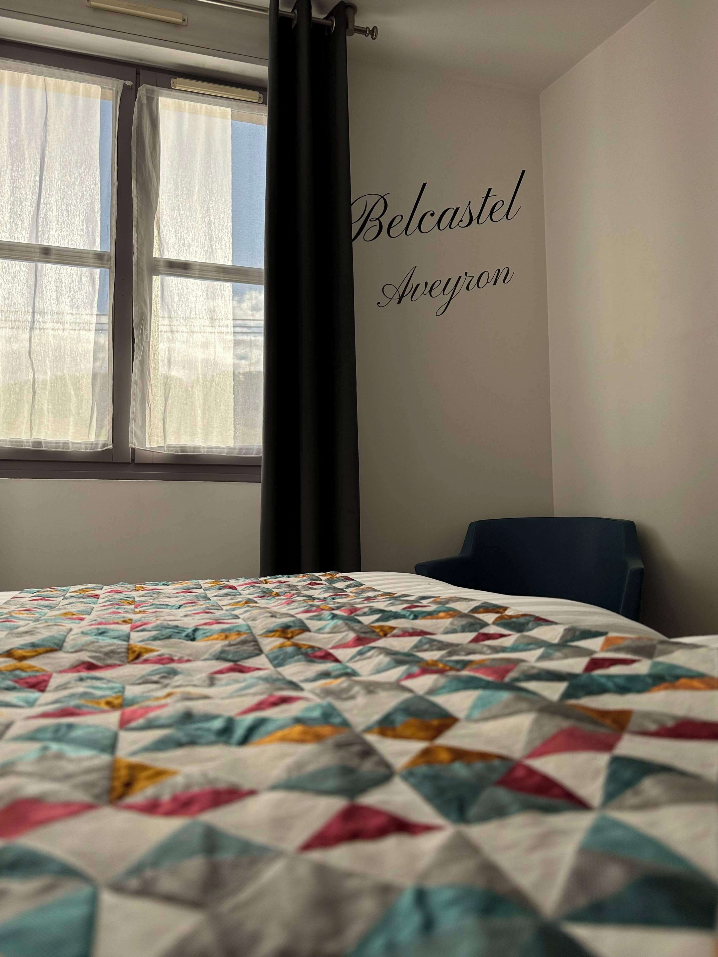 A room with a bed, a chair, a window with curtains, and a wall with the text 'Belcastel Aveyron'.