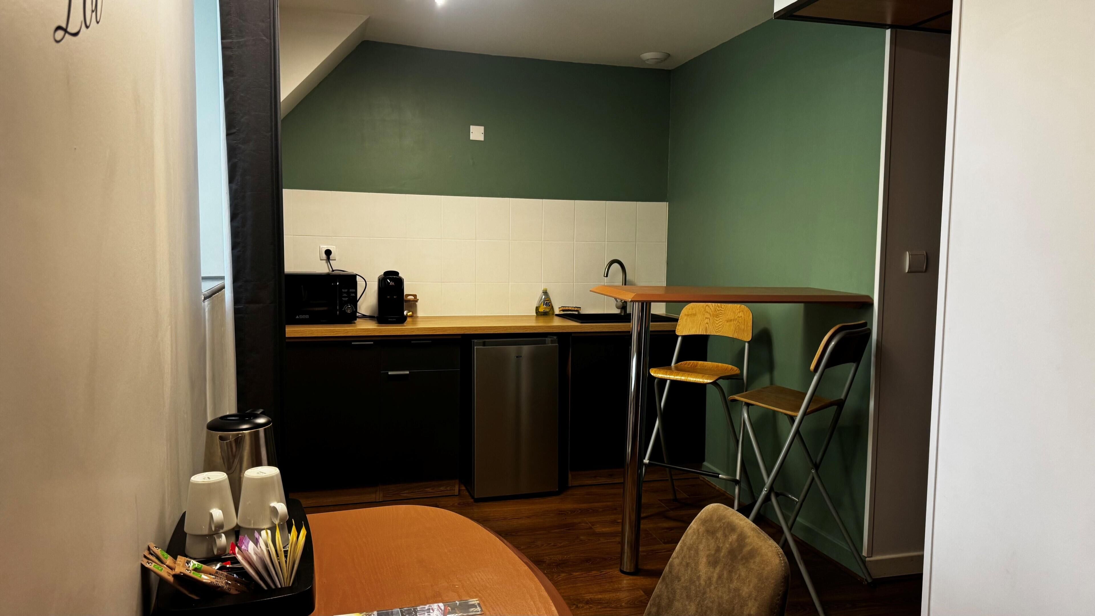 A compact kitchen with a dishwasher, a coffee maker, and a sink. There is a dining table with chairs and a small coffee station with cups and utensils.