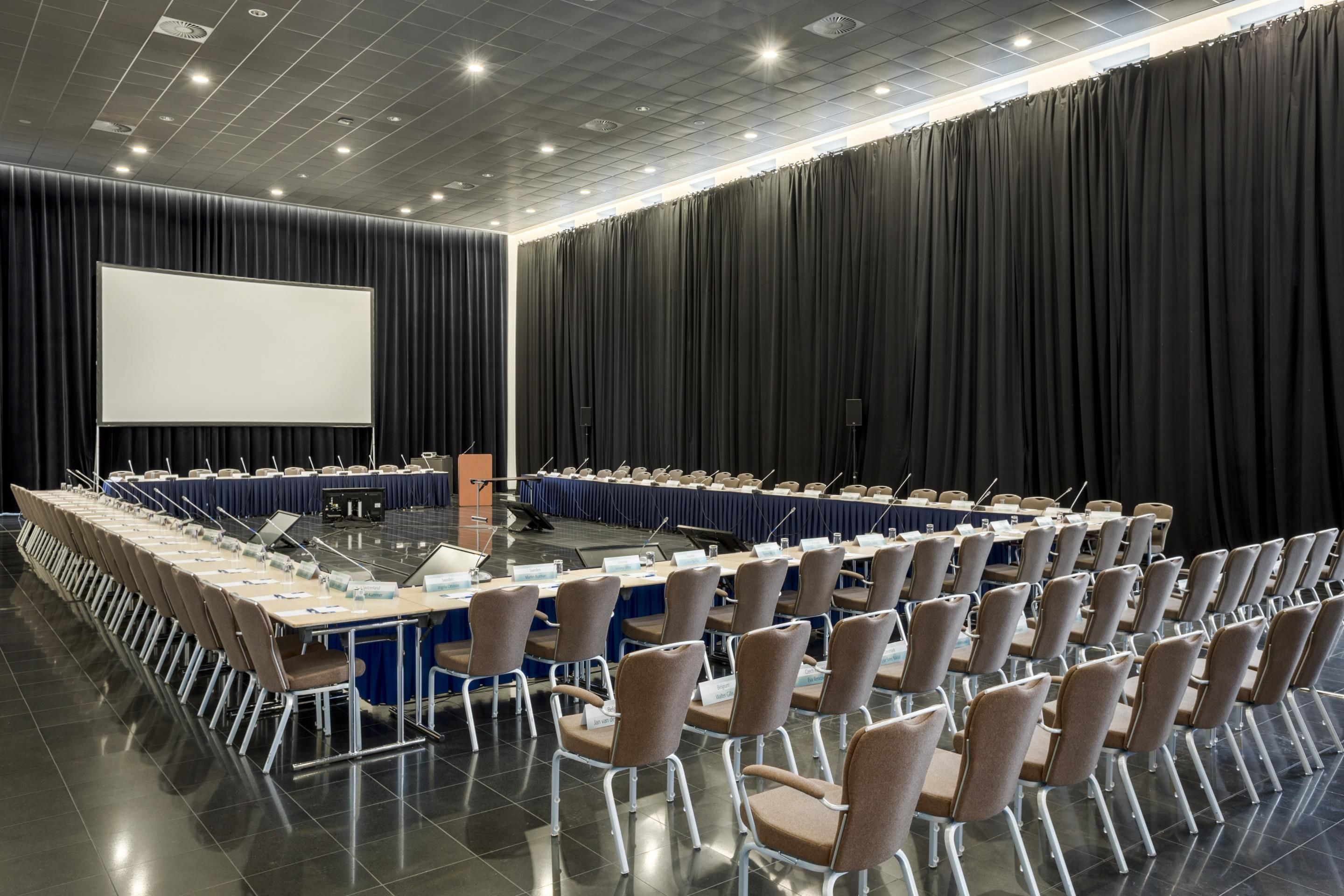 Un salón de conferencias con una larga mesa, sillas, una pantalla de proyector y un telón oscuro de fondo.