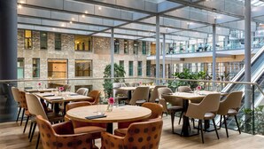A modern indoor dining area with round tables and chairs, large windows, and an adjacent staircase. - NH Den Haag (The Hague)