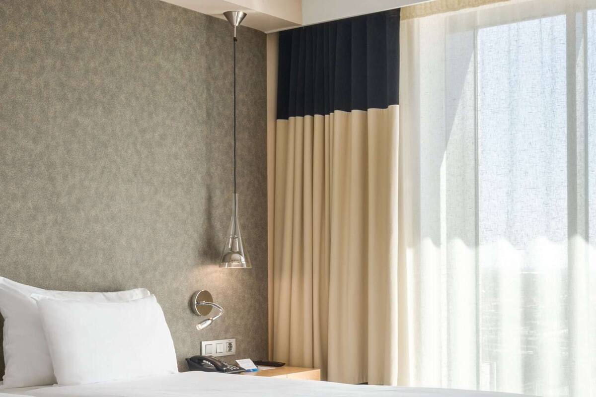A hotel room with a bed, a bedside table with a lamp, a phone, and a window with curtains.