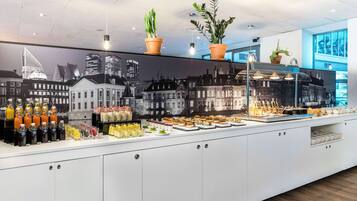 Un bufé con diversas opciones de comida y bebidas, plantas en macetas y un mural con vista a la ciudad.