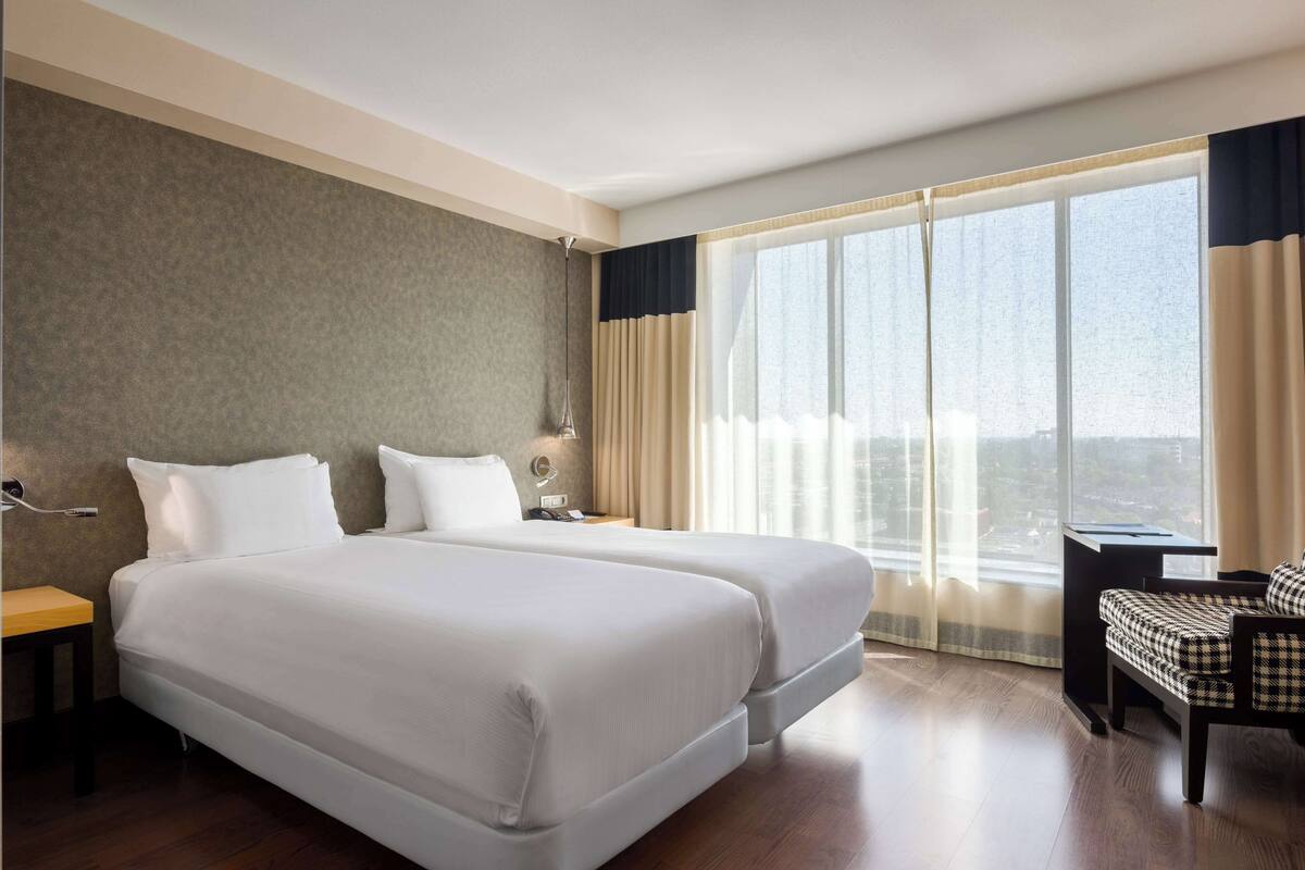 A hotel room with two beds, a desk, a chair, and a large window with curtains.