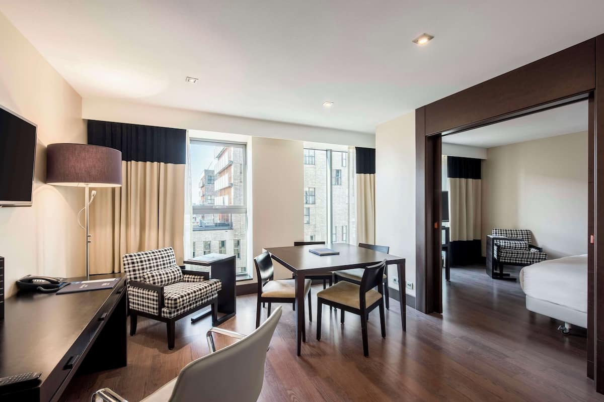 A hotel room with a desk, two chairs, a bed, and a view of buildings through large windows.