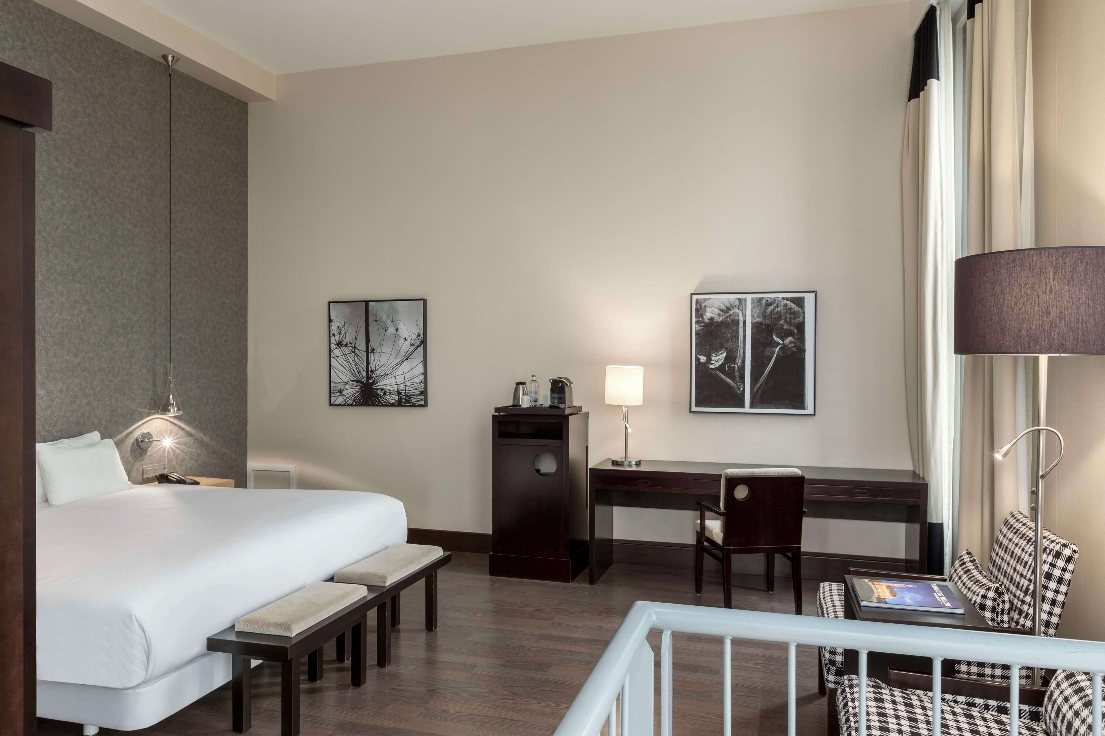 Habitación de hotel con cama, escritorio, silla, lámpara, obras de arte y vistas al exterior.