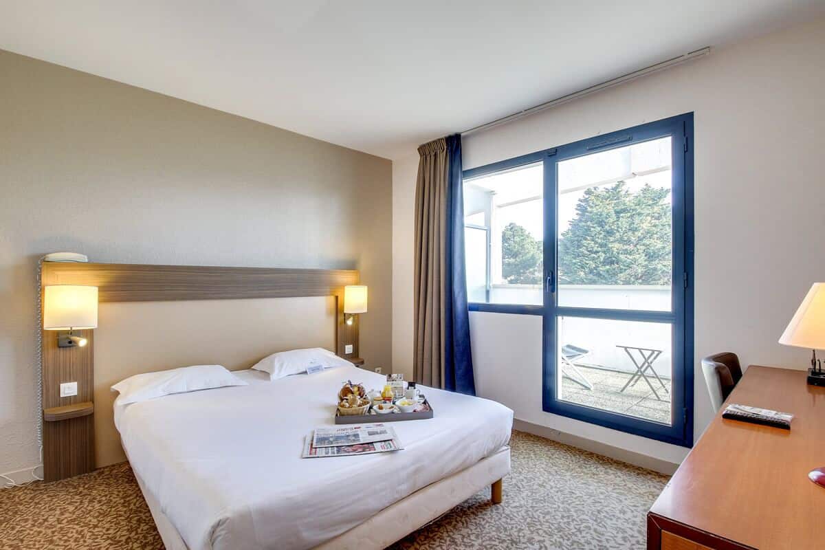 A hotel room with a bed, a desk, a window with curtains, and a view of the outdoors.