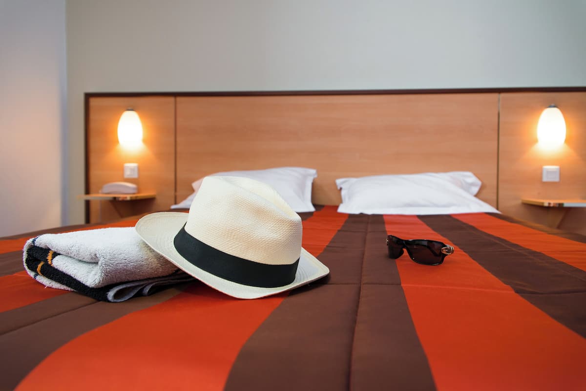 A neatly made bed with a striped bedspread, a folded towel, a hat, and a pair of sunglasses on the bed.