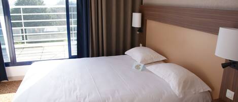 A hotel room with a large bed, a nightstand, a wall-mounted lamp, and a window with curtains.