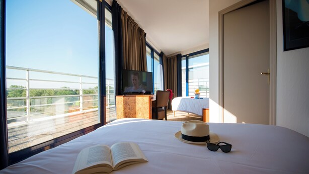 Una habitación de hotel con una cama grande, un libro abierto, un sombrero y gafas de sol sobre la cama. Hay un televisor en un mueble de madera, una silla y una mesa con mantel blanco. La habitación tiene ventanas amplias con cortinas, y se ve una puerta.