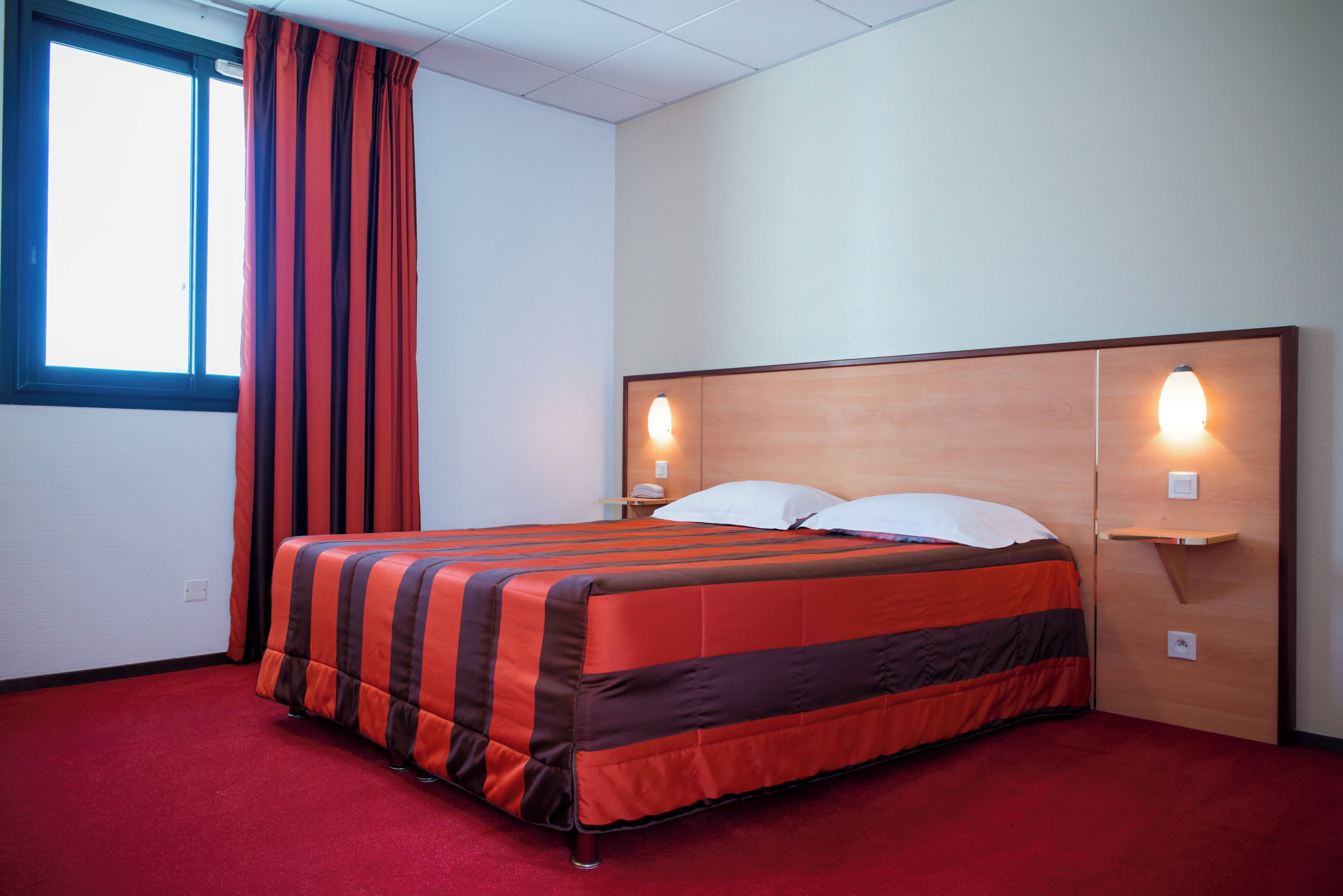 Ein Hotelzimmer mit Bett, rotem Teppich, Vorhängen, einem Fenster und Wandleuchten.