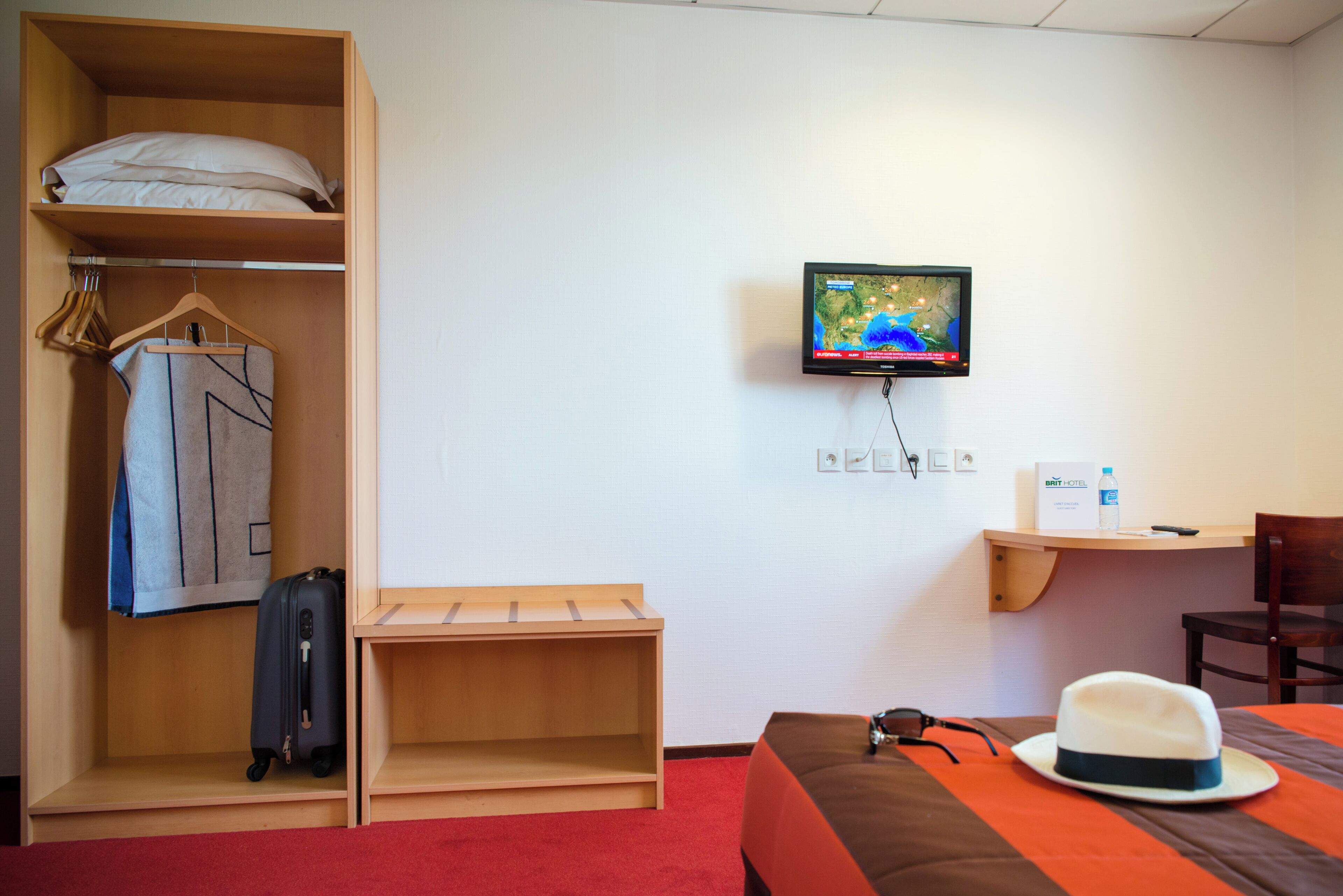 Une chambre d’hôtel avec un lit, un téléviseur fixé au mur, une armoire en bois, un bureau avec une chaise et une moquette rouge.