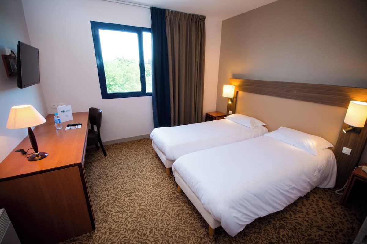 A hotel room with two beds, a desk with a lamp, a chair, a window with curtains, and a wall-mounted TV.