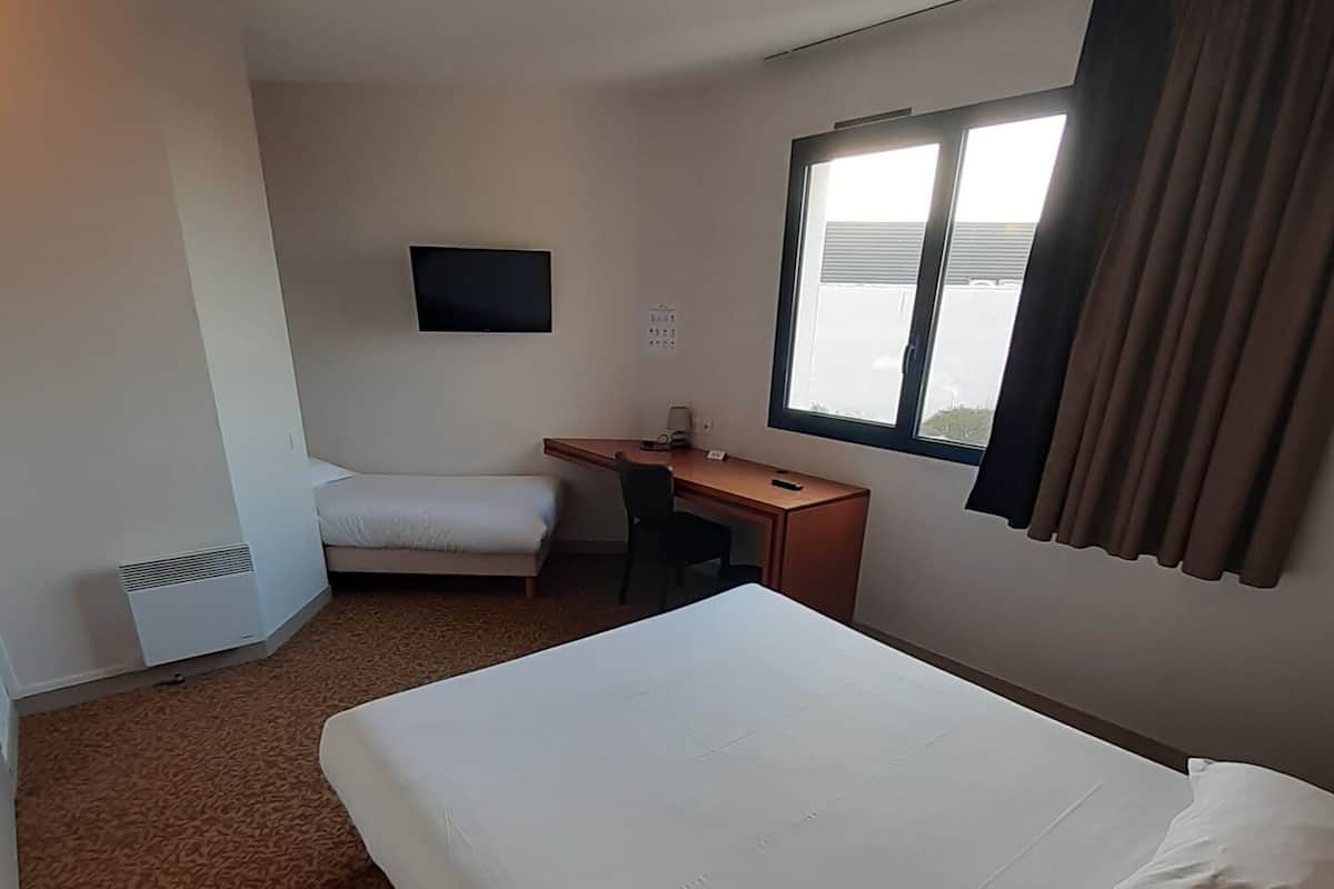 A hotel room with a large bed, a desk, a chair, a flat-screen TV mounted on the wall, and a window with curtains.