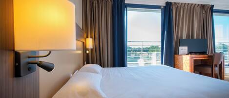 Quarto de hotel com uma cama grande, uma secretária com computador, uma cadeira e vista para a cidade pela janela.