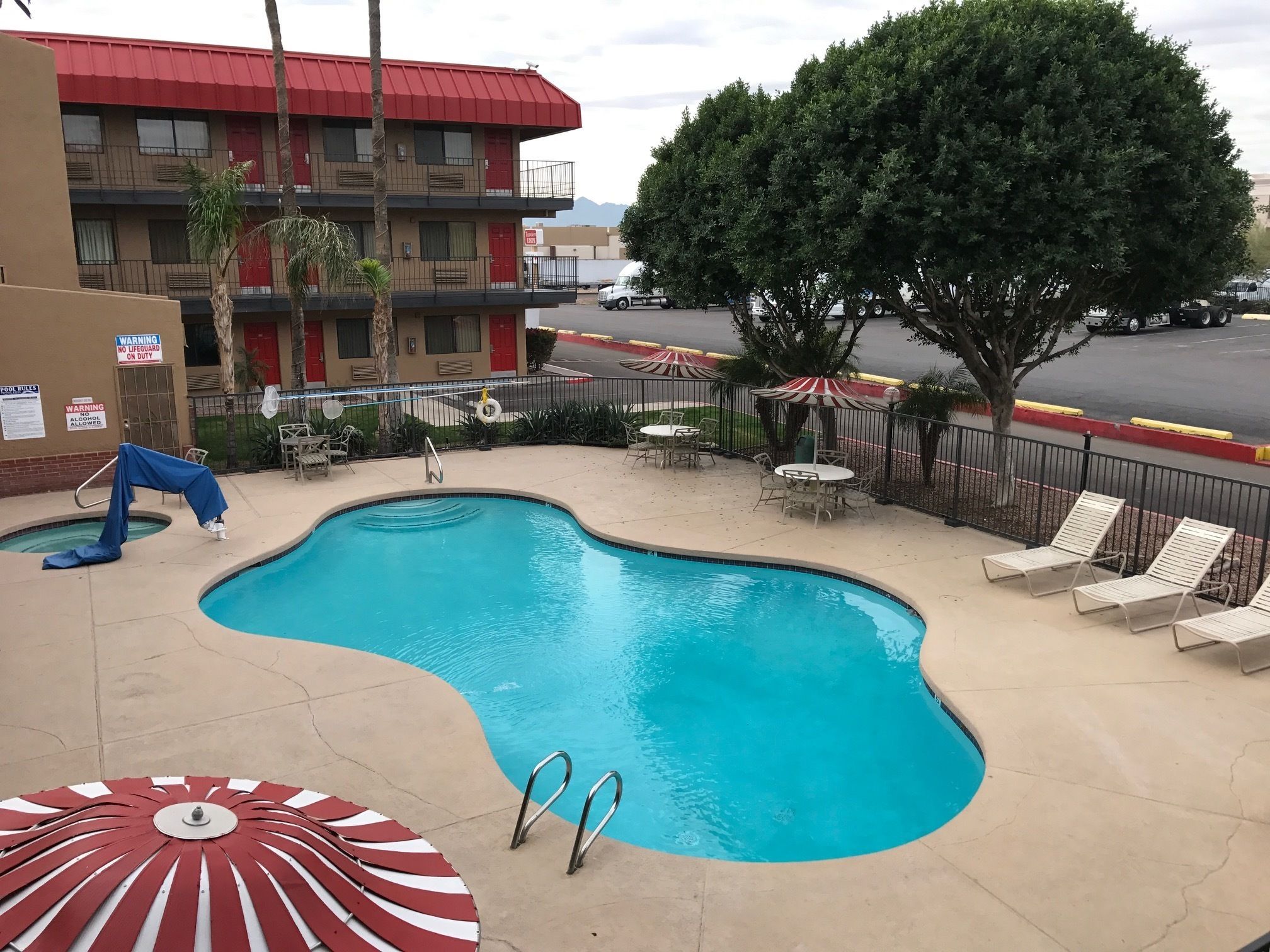 Piscina com espreguiçadeiras e guarda-sóis, área cercada com mesas e cadeiras, e um edifício com telhado vermelho e sacadas.