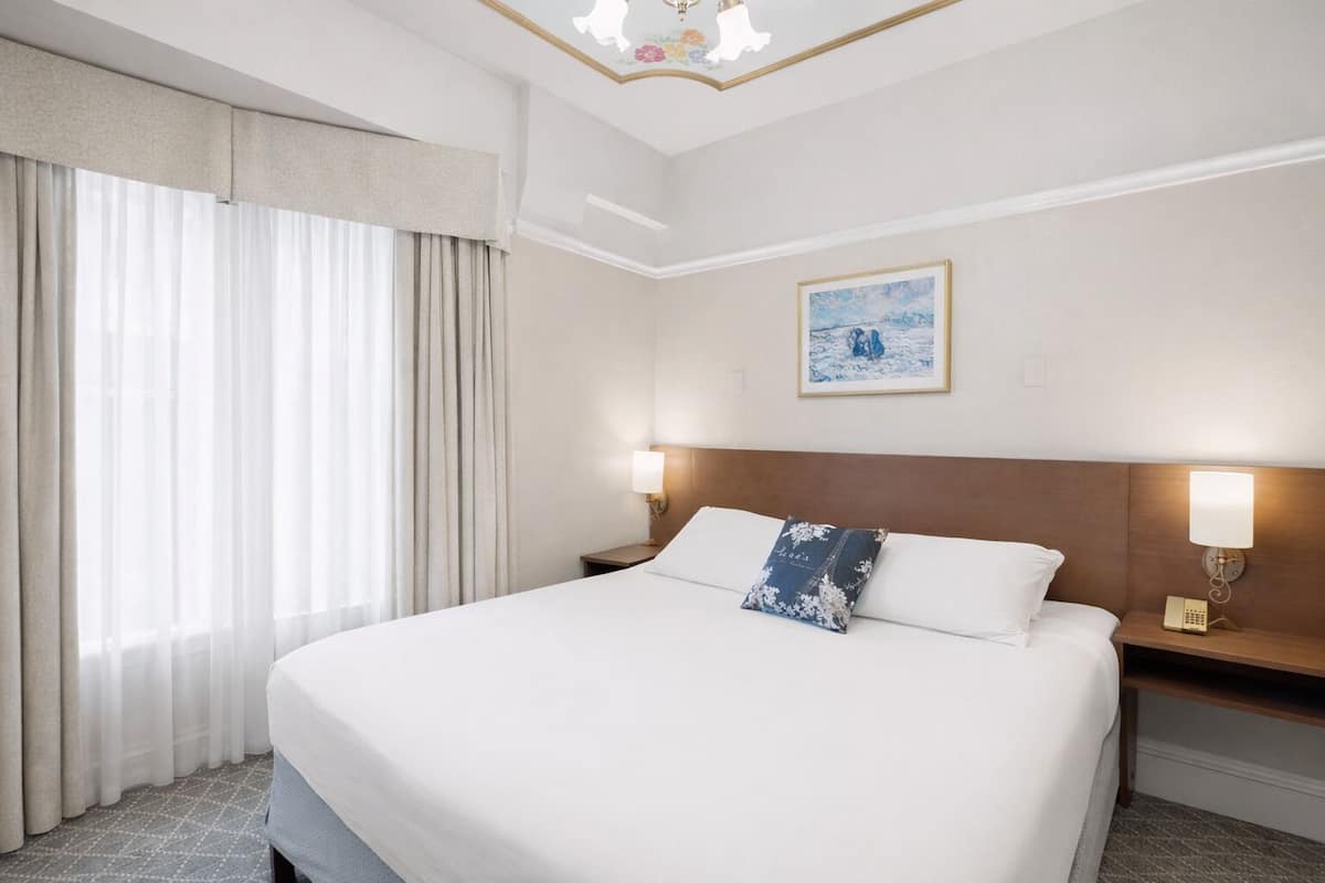 Une chambre d’hôtel avec un grand lit, deux tables de chevet avec des lampes, un tableau au mur et une fenêtre avec des rideaux.