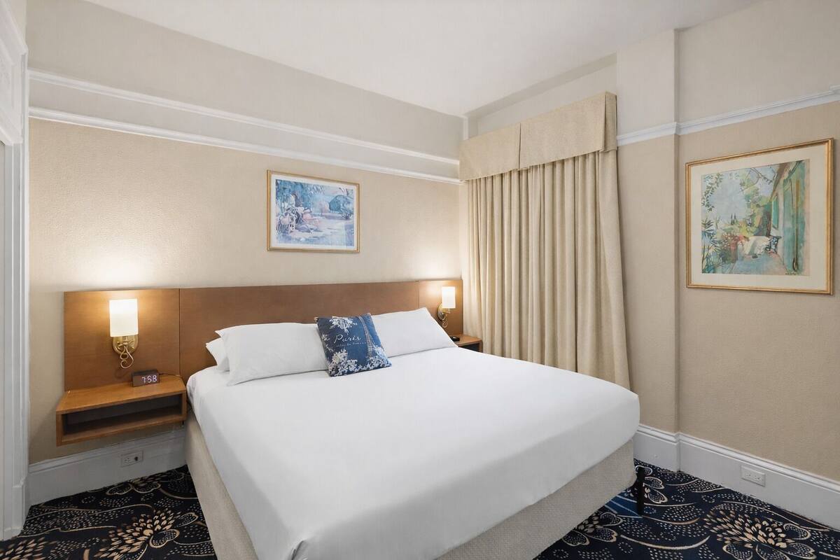 Une chambre d’hôtel avec un grand lit, deux tables de chevet, une petite table, une horloge murale et deux œuvres d’art encadrées.