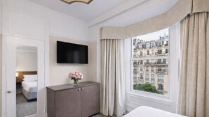 A hotel room with a bed, a TV mounted on the wall, a cabinet with a vase of flowers, and a window with curtains. - Cornell Hotel (San Francisco)