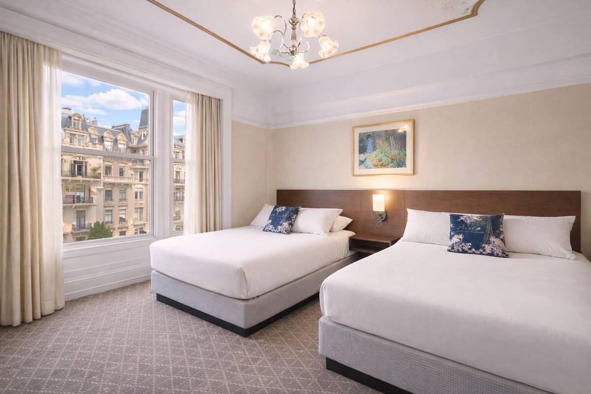 Une chambre d’hôtel avec deux lits, une grande fenêtre, un lustre et un tableau au mur.