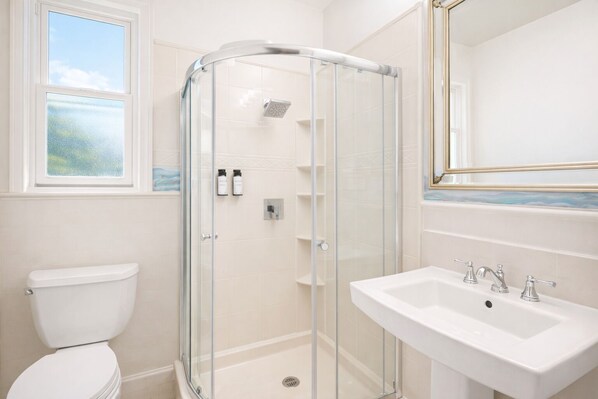 A bathroom with a glass-enclosed shower, a toilet, and a sink with a mirror above. - Cornell Hotel (San Francisco)