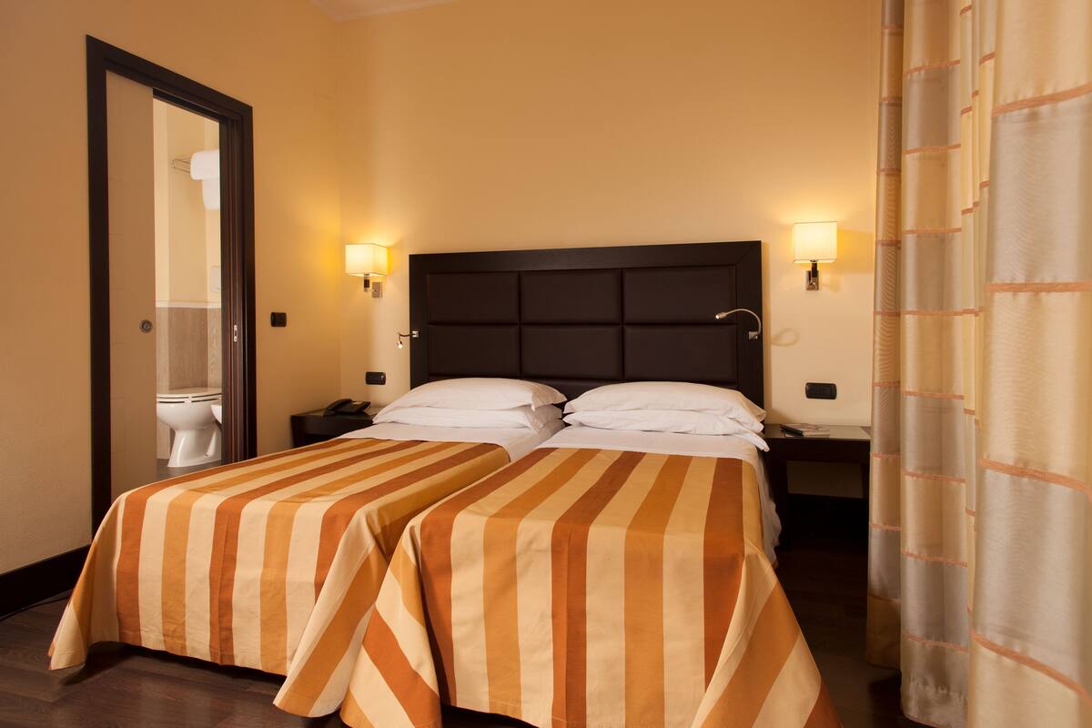 A hotel room with two beds, a dark headboard, bedside lamps, and a bathroom visible through an open door.