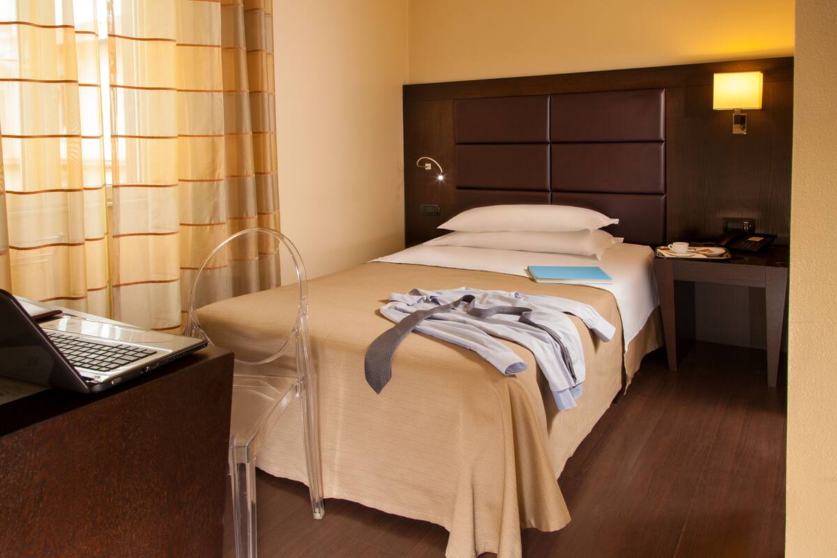 A hotel room with a bed, a desk with a laptop, a bedside table with a phone, and a window with curtains.