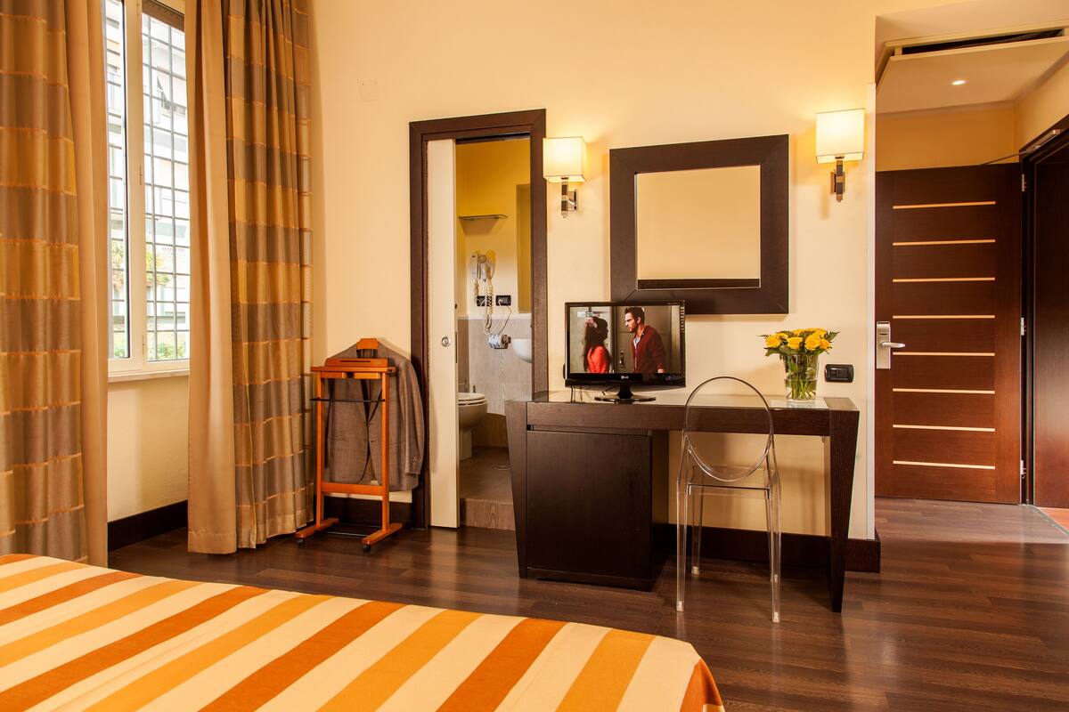 A hotel room with a bed, a desk with a computer, a mirror, and a bathroom visible through an open door.