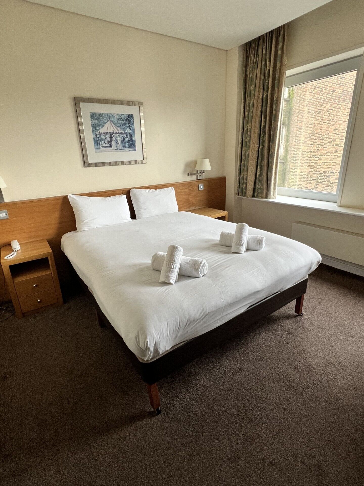 A hotel room with a large bed, a nightstand, a lamp, a framed picture on the wall, and a window with curtains.