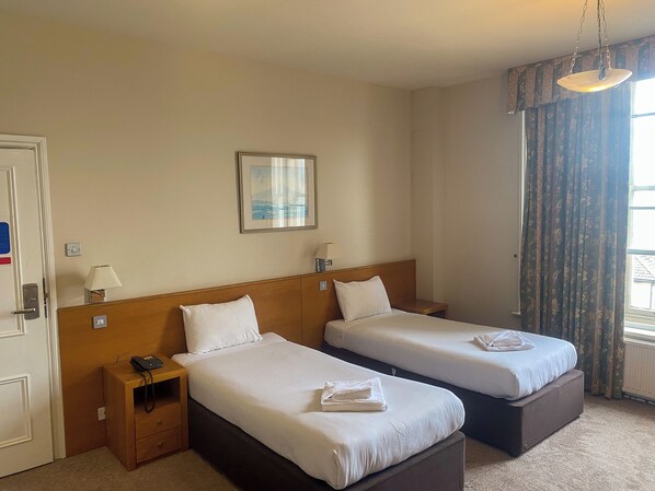 A hotel room with two beds, a bedside table with a lamp, a framed picture on the wall, and a window with curtains. - Cavendish Hotel (Eastbourne)