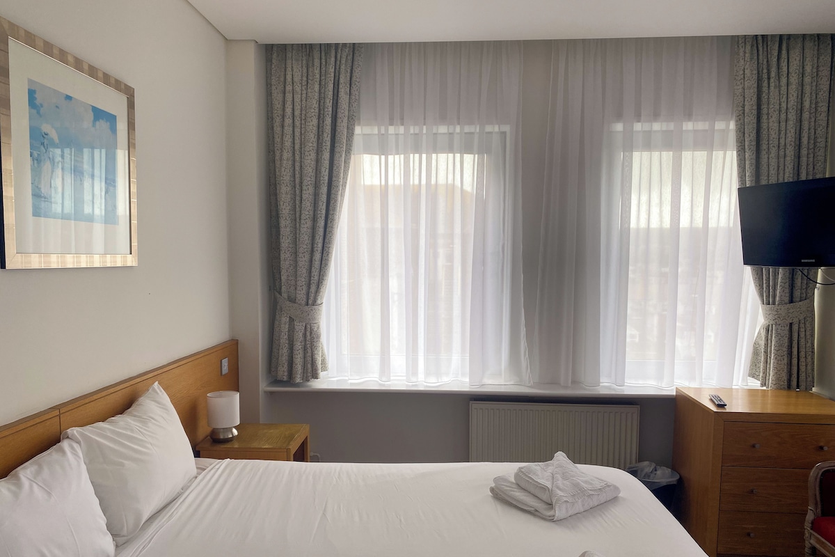 A hotel room with a large bed, a wooden headboard, a bedside table with a lamp, a dresser, a television, and a window with sheer curtains.