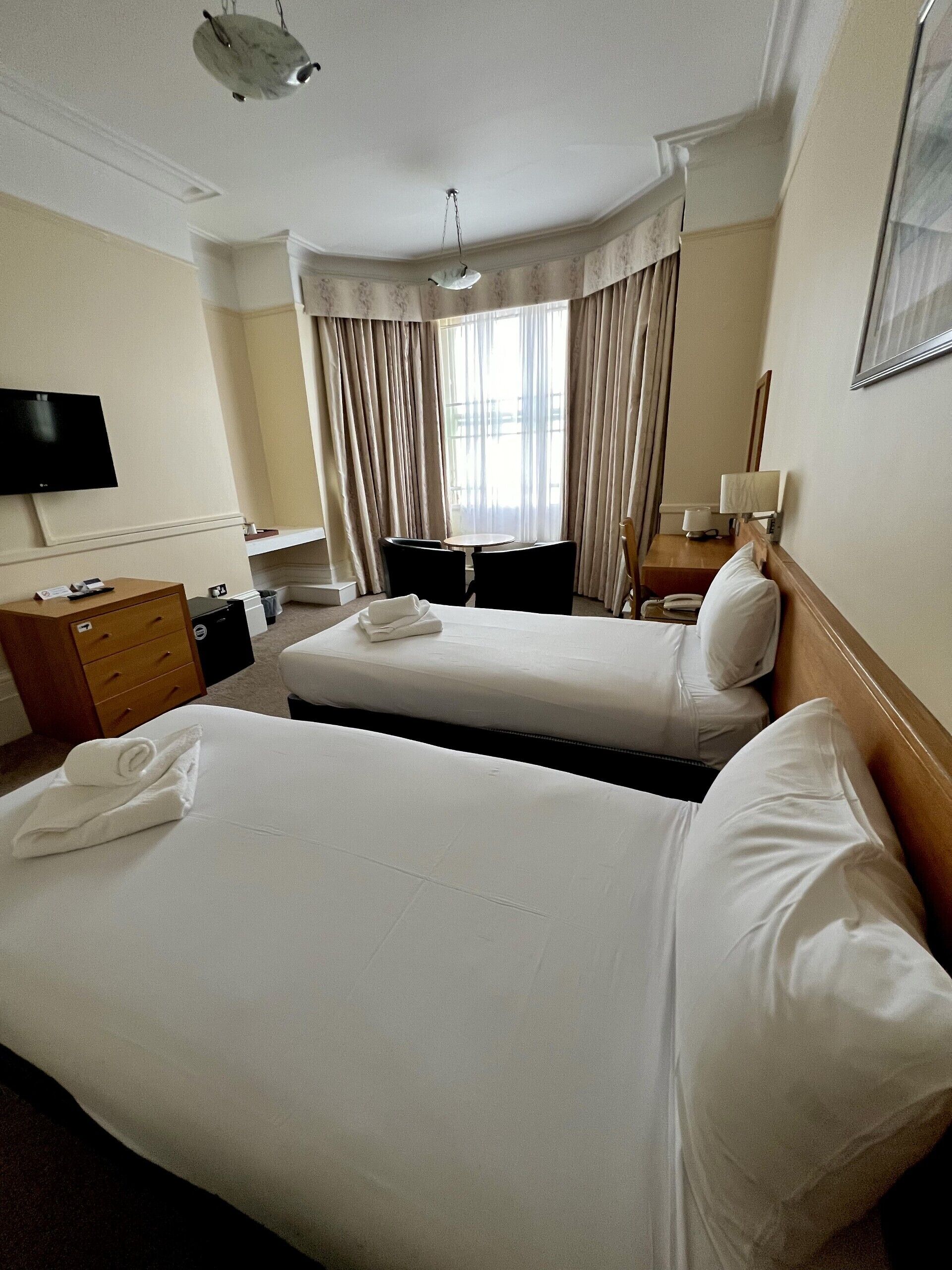 A hotel room with two beds, a desk with a computer, a TV, and a window with curtains.