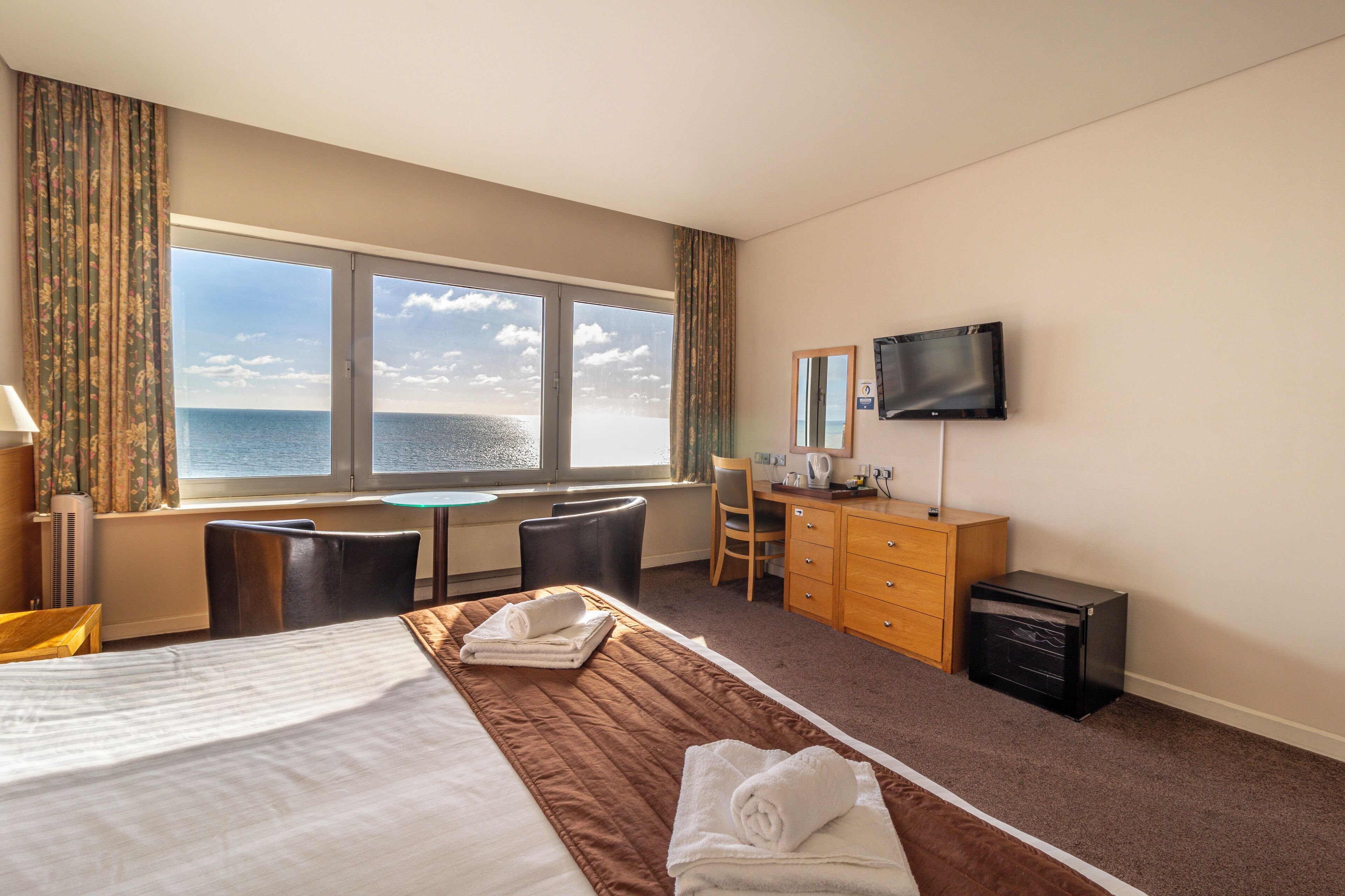 A hotel room with a large bed, a desk, a chair, a TV, and a view of the sea.