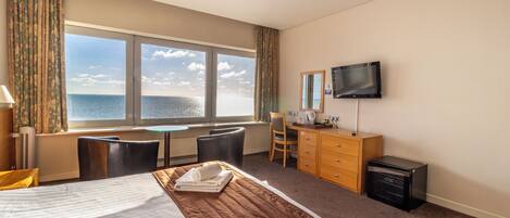 A hotel room with a large bed, a desk, a chair, a TV, and a view of the sea.