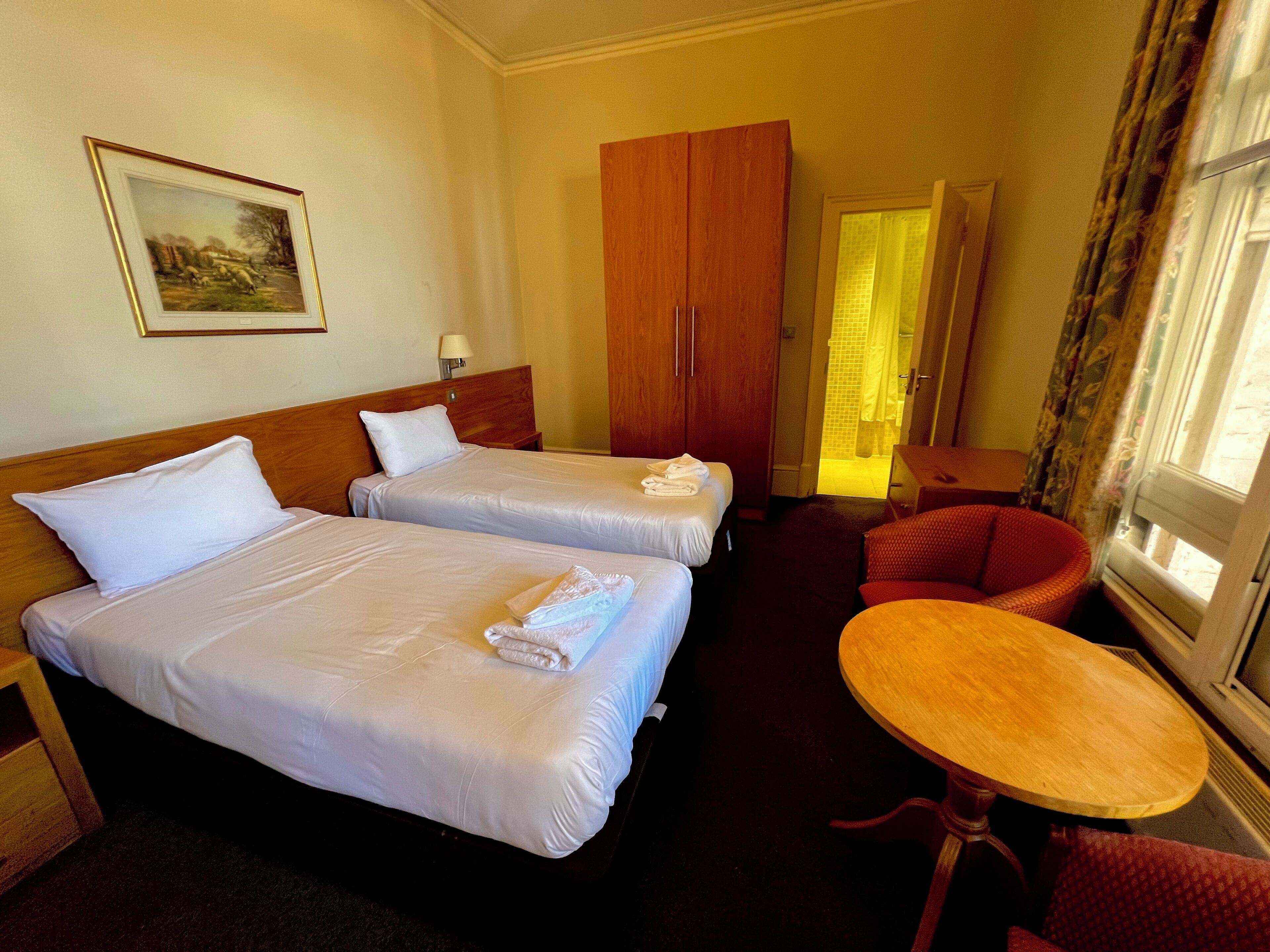 A hotel room with two beds, a wooden wardrobe, a small table, and a chair.