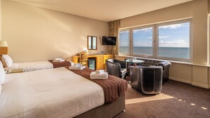 A hotel room with two beds, a desk with a computer, a TV, and a large window with a view of the sea. - Cavendish Hotel (Eastbourne)
