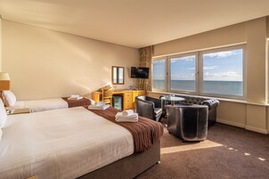 Ein Hotelzimmer mit zwei Betten, einem Schreibtisch mit Computer, einem Fernseher und einem großen Fenster mit Meerblick.