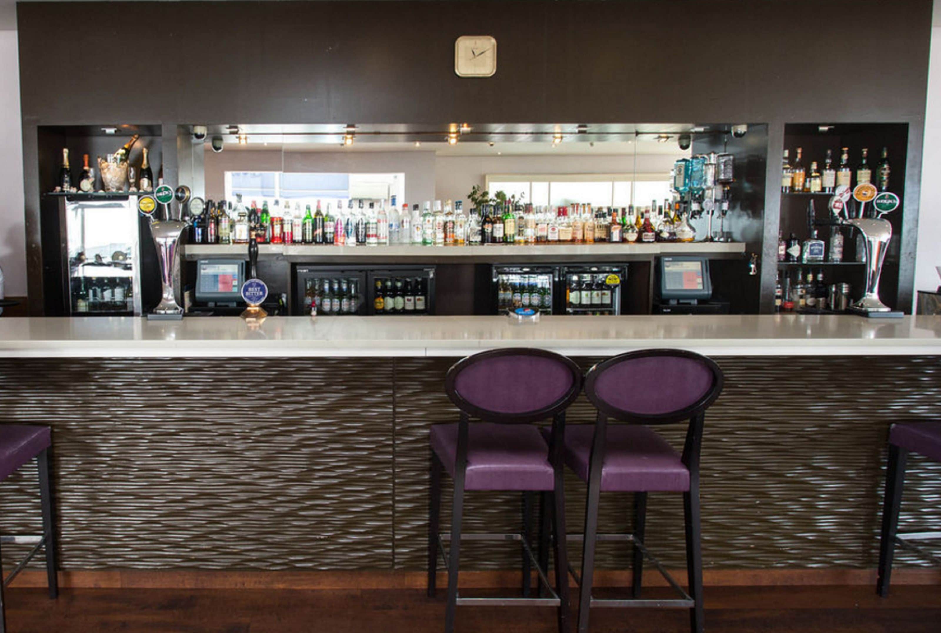Un espace bar avec un comptoir, de nombreuses bouteilles d’alcool et deux tabourets de bar violets.